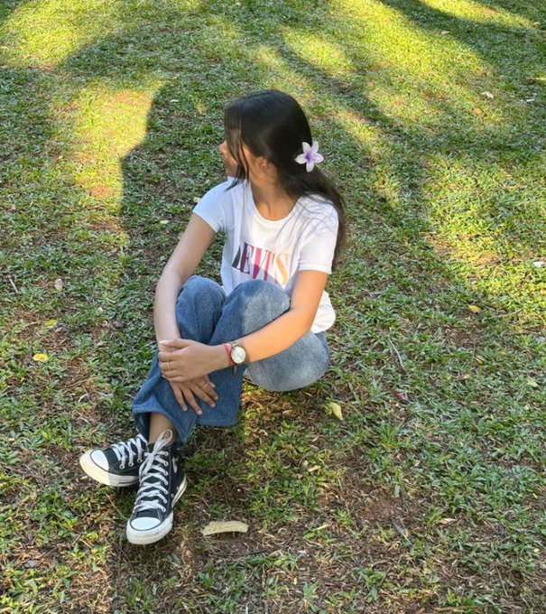 A photograph of a young woman sitting cross-legged on a grassy field, wearing a white 'LEVI'S' t-shirt, blue jeans, and black Converse sneakers, with a small purple flower tucked behind her ear.