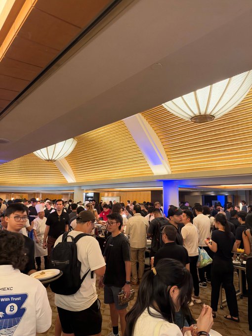 A crowded indoor event space filled with attendees mingling, with one person's shirt visibly displaying "MEXC" and "With Trust" text. The post critiques Solana, suggesting the large gathering is a façade—claiming 99.9% of Solana DEX volume is bot-driven rather than genuine community engagement.