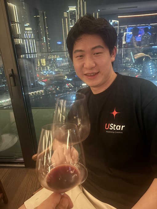 A young man wearing a black "uStar - Powered by Creators" t-shirt smiles while holding two wine glasses against a stunning nighttime city skyline backdrop. The setting appears to be a high-rise dinner gathering with someone clinking glasses in celebration.