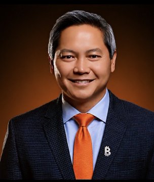 A professional portrait of a smiling middle-aged man in a dark suit, light blue shirt, and orange tie with a "B" lapel pin.