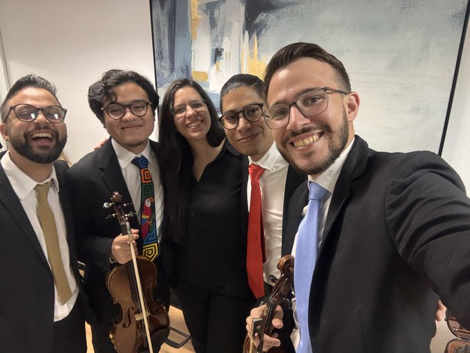 A group of five smiling people in formal attire, with two holding string instruments (violin and viola), taking a selfie indoors.