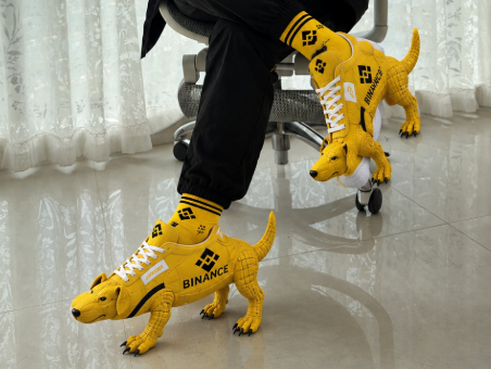 A person wearing novelty yellow LEGO sneaker-shaped footwear featuring the Binance logo, standing on a tiled floor.