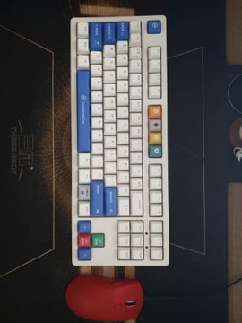 A white mechanical keyboard with blue accent keys sits on a black mousepad, accompanied by a red earbuds case.
