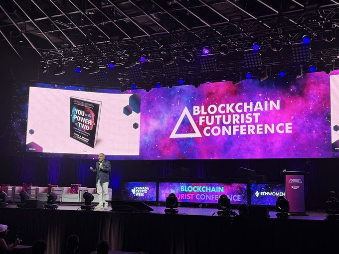 A man speaks on stage at the Blockchain Futurist Conference, with large screens displaying the event title and a featured book cover "You Power -TWO-." The image captures the opening keynote moment, aligning with the post about Don Tapscott's talk on rebuilding Canada for the new technology order.