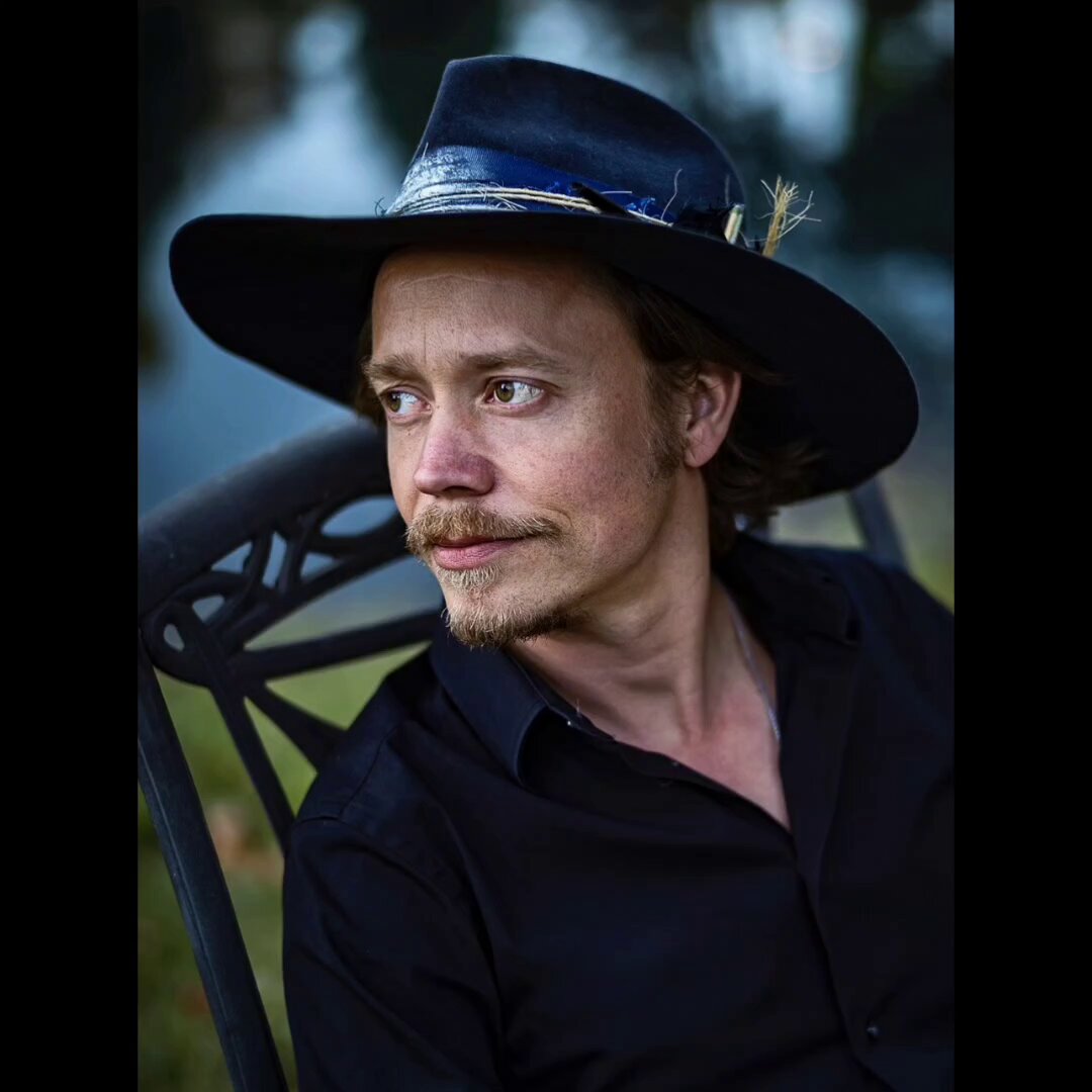 The image features a man wearing a black hat sitting in a chair outdoors. The photo has been edited to include text that says "Many people believe that the Iran war is about energy dominance. It really seems to be about restructuring the monetary system from the petrodollar with crypto stablecoin. USDT (Tether) found it beginnings with the likes of Brock Pierce, Howard Lutnick, Canton Fitzgerald, and". The man in the hat appears to be looking off into the distance or at something else outside the frame, possibly related to the post about cryptocurrency and the Iran war.