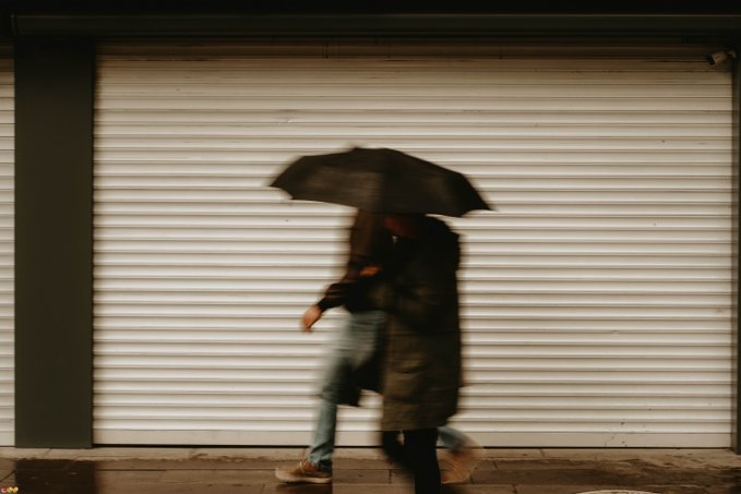 Blurred motion photo of two people walking under a dark umbrella past a white corrugated metal storefront shutter.