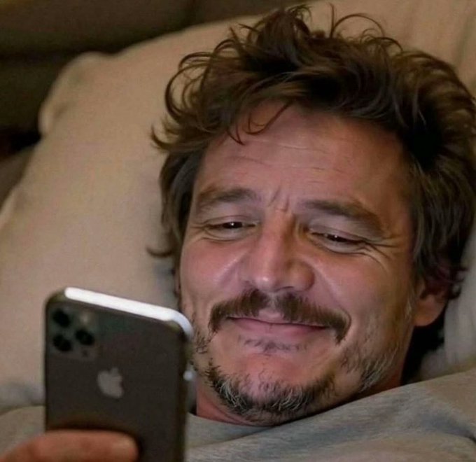 A man, resembling actor Pedro Pascal, lies in bed smiling while looking at an iPhone (visible Apple logo). The image playfully illustrates the post's theme of resorting to a phone for work-related tasks, such as checking charts, due to not having a laptop.