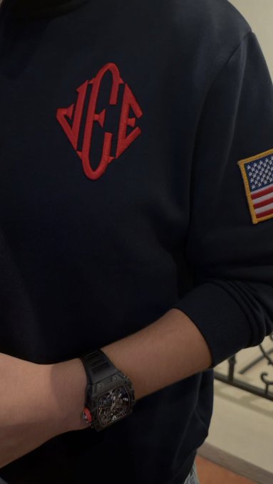 A person wearing a navy sweatshirt featuring a red diamond-shaped "ACE" logo on the chest and an American flag patch on the sleeve, while wearing a large black sports watch.