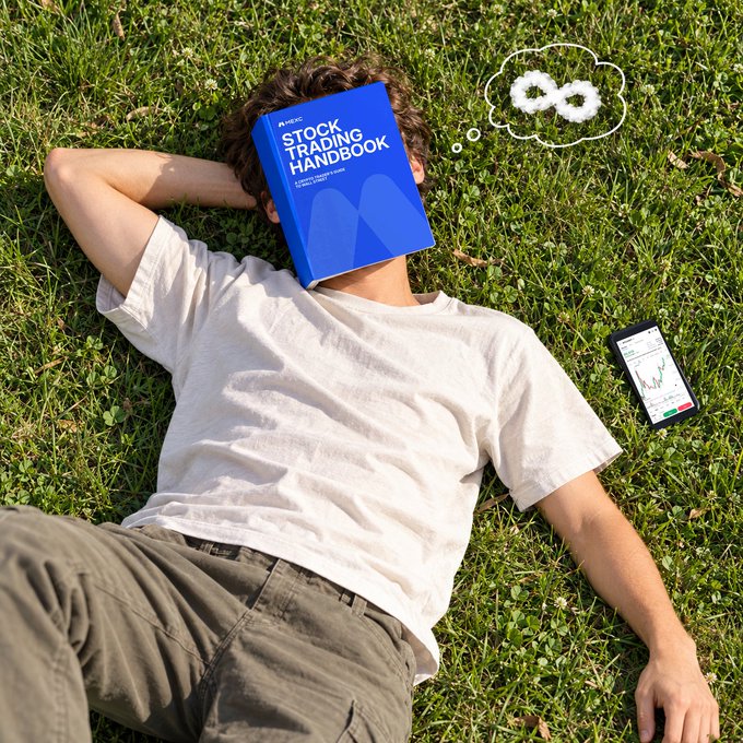 A man lies on his back in the grass with a blue "Stock Trading Handbook" covering his face, while his phone next to him displays a stock chart. A thought bubble above him shows an infinity symbol, playfully suggesting he's stuck in an endless cycle of "doing your own research" instead of truly relaxing outdoors.