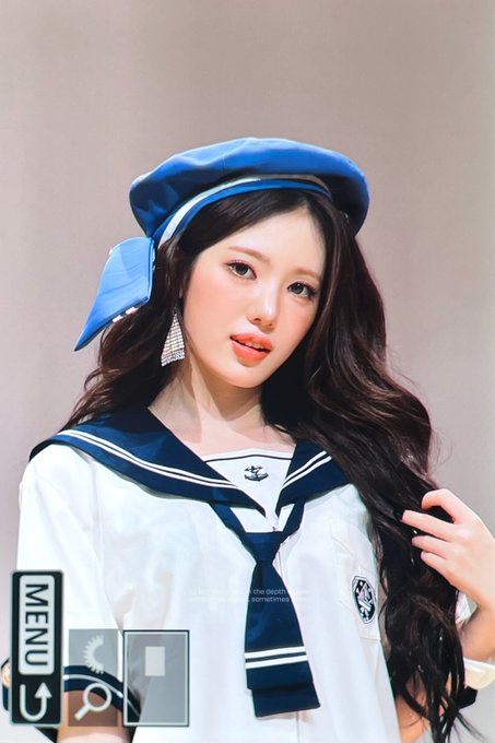 A woman with long dark hair wears a blue and white sailor-style outfit and a matching blue beret, with subtle text reading "i am the deer that is born anew" visible on the image.