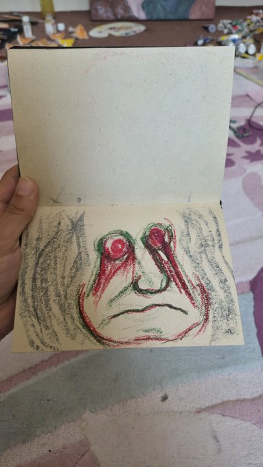 A child's crayon drawing in an open notebook showing a colorful, abstract face with large eyes and a frowning mouth.