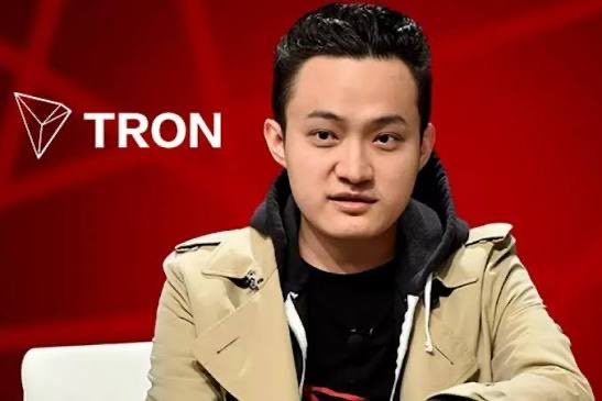 A young man wearing a tan trench coat over a dark hoodie is shown against a red background featuring the TRON logo.