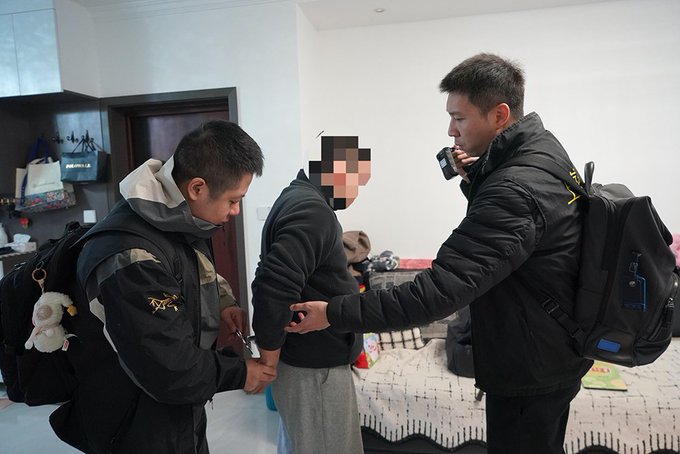Two officers, one handcuffing a suspect whose face has been pixelated for privacy, while the other speaks into a two-way radio during an arrest in a residential bedroom. The image depicts law enforcement action related to the reported Shanghai police investigation into a major underground banking case involving cryptocurrency-based cross-border currency exchange, with over 200 billion yuan implicated and more than 70 suspects arrested.