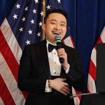 A smiling man in a tuxedo speaks into a microphone in front of an American flag backdrop. The image likely depicts Justin Sun, founder of TRON, who is being referenced in the post discussing Bitcoin's evolution into a global macro asset and its influence on his decision to build TRON.