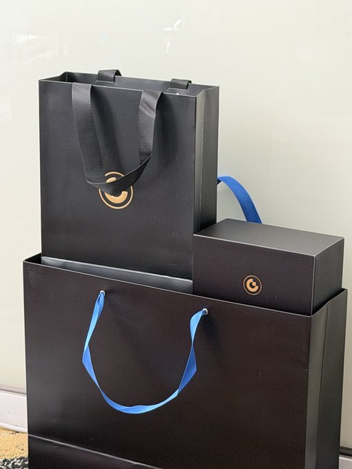 Two black gift bags with blue and black ribbon handles, featuring a small gold circular logo, are displayed alongside a matching black gift box with the same logo. The image relates to the post by showing the three gift boxes the user applied for after earning 2M USD1 through farming on Gate.