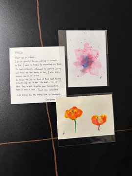A handwritten thank-you note sits next to two small watercolor paintings of flowers on a dark surface.