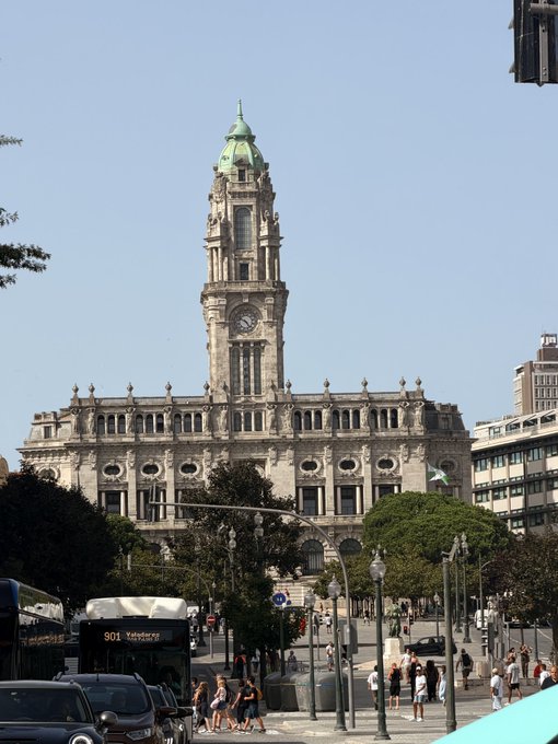 A historic ornate building with a tall clock tower and green dome, with pedestrians walking on the street below under a clear blue sky.
