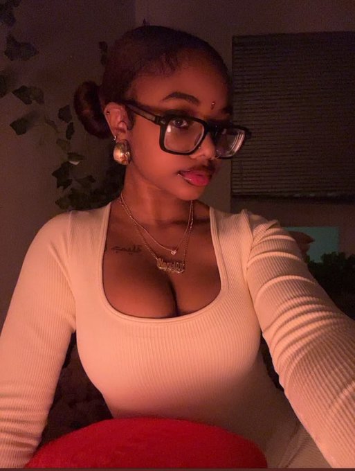 A woman poses for a mirror selfie wearing black-framed glasses, gold hoop earrings, and layered nameplate necklaces over a cream-colored ribbed top, with a small tattoo visible on her chest, matching the relaxed and positive vibe of wishing her followers a productive new week.