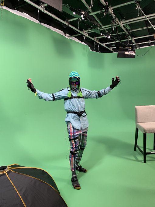 A person in a motion-capture suit with green hair, patterned pants, and reflective sunglasses stands with arms raised in front of a green screen in a studio.