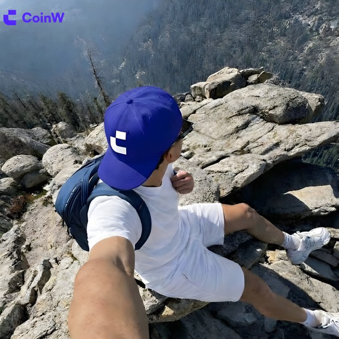 A hiker wearing a blue "CoinW" cap and white outfit sits perched on a rocky mountain ledge, dramatically stretching their arm out to capture a selfie that emphasizes the dizzying drop below—taking the "zoom out" advice literally with a breathtaking cliffside perspective.