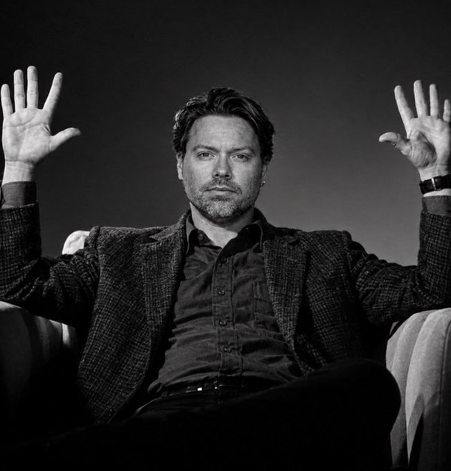 A black-and-white photograph captures a bearded man in a dark blazer and shirt sitting in a chair with both hands raised palms-out in a shrug of exasperation or disbelief. The image visually mirrors the post's tone of weary resignation, illustrating the frustration of the HEX community enduring years of delays, legal battles, and skepticism.