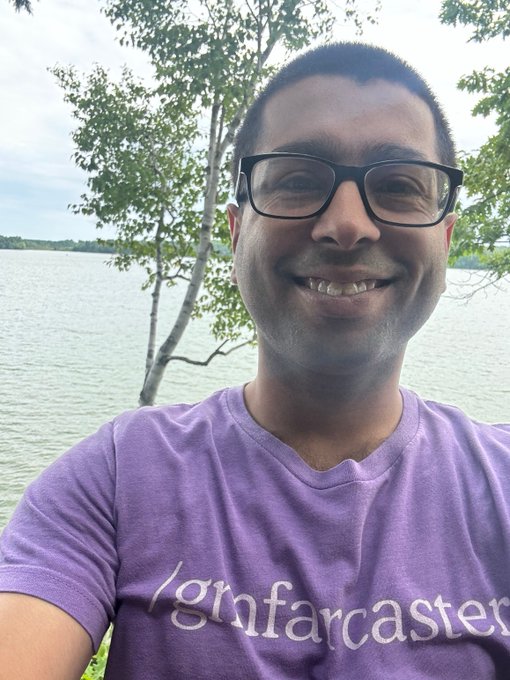 A smiling man with glasses wearing a purple t-shirt that reads "/gm farcaster" poses for a selfie in front of a lake with trees.