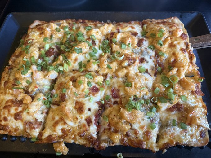 A rectangular sheet pan pizza topped with melted cheese, bacon pieces, and chopped green onions, cut into squares and resting on a black baking tray.