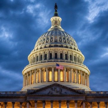The illuminated dome of the U.S. Capitol building at dusk against a dramatic blue cloudy sky, with an American flag visible.