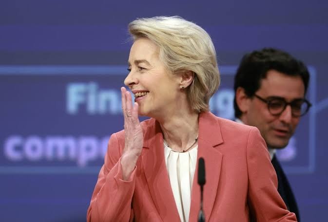 Ursula von der Leyen smiling and waving while wearing a coral blazer, standing at a podium with partial background text 'Final' and 'competi' visible.