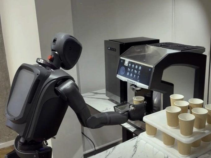 A humanoid robot operates a coffee machine in a kitchen, with a tray of empty cups waiting to be filled.
