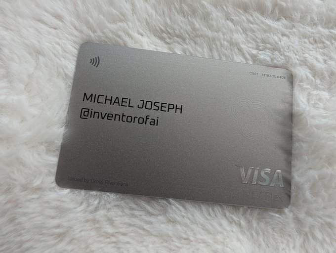 Photo of a gray metal Visa Infinite Flex credit card issued by Cyprus Popular Bank, engraved with the name 'MICHAEL JOSEPH' and the handle '@inventorofai', resting on white fluffy fabric.