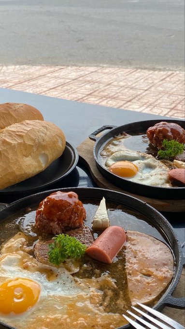 Two cast iron skillets contain hearty meals with sunny-side-up eggs, sausages, meat patties, and garnishes, served alongside crusty bread rolls on a sidewalk table.