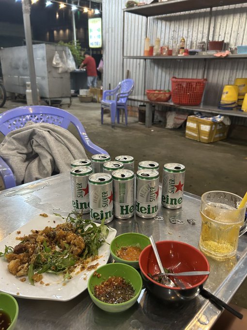An outdoor evening street food scene featuring a metal table with eight Heineken Silver beer cans, a plate of grilled meat with herbs, small bowls of dipping sauces, and a glass of beer, set against a backdrop of plastic chairs and a food stall.