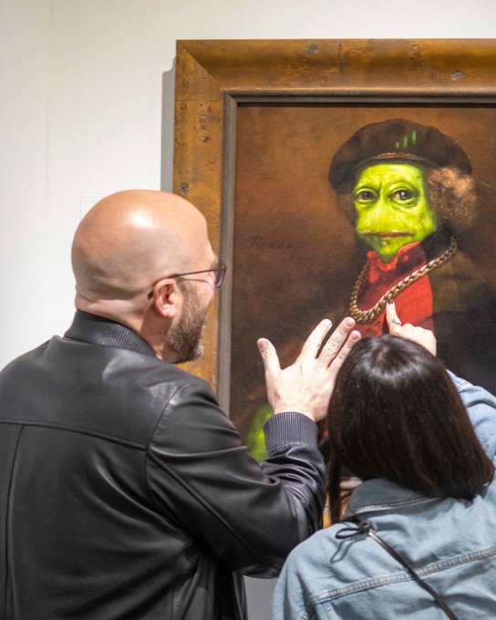 A bald man with glasses and a beard in a black leather jacket stands before a framed portrait in which the subject's face has been digitally altered to green Shrek-like skin with glowing eyes.