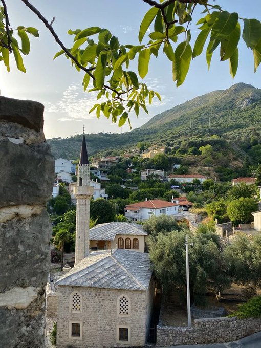 A scenic view of a traditional stone mosque with a tall minaret in a Mediterranean village, framed by tree branches with green leaves in the foreground, surrounded by hills and red-roofed houses.