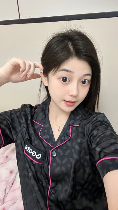 A selfie of a young woman with long dark hair wearing a black 'Paris' patterned pajama top with pink trim, posing indoors.