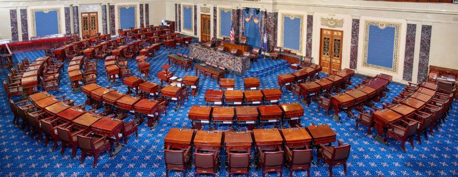 An overhead view of an empty legislative chamber with rows of wooden desks and chairs arranged in a semicircle on a blue patterned carpet.