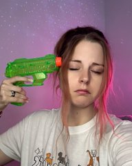 In the image, a woman with pink hair is wearing a white t-shirt that features an image of dogs printed on it. She has her head covered by a green plastic toy gun held behind her ear, which appears to be aimed at her head, suggesting she may have been playing around or experimenting with the toy in a playful manner. The photo also contains some text, which includes "ETH holders rn $ETH goes below !" and "still acting calm". The context of this image is not explicitly clear, but it seems that the woman's interest in the toy gun might be related to an event or situation involving the ethical holder of those particular items.