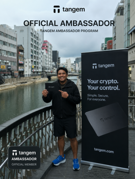A man stands on a bridge holding a Tangem card, with text reading 'OFFICIAL AMBASSADOR' and 'TANGEM AMBASSADOR PROGRAM,' alongside a banner stating 'Your crypto. Your control. Simple. Secure. For everyone. tangem.com.'