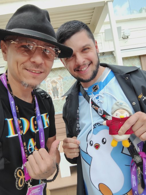Two smiling men pose together at what appears to be a convention, one wearing a black fedora and sunglasses with an 'Anime' badge, the other in a Pokémon hoodie holding a small plush toy.