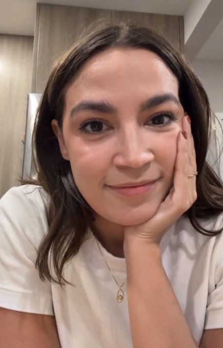 A selfie of a smiling young woman with brown hair wearing a white shirt and delicate gold necklace, resting her face on her hand.
