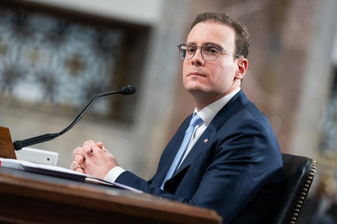 A man in a dark suit, light blue tie, and glasses sits at a witness table with a microphone in front of him, looking upward in what appears to be a Senate hearing room.