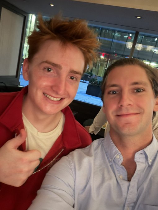 A selfie of two young men, one in a red jacket with messy brown hair smiling widely, and the other in a light blue button-down shirt smiling slightly.