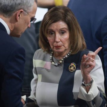 Nancy Pelosi, wearing a pin with "Pelosi" visible on her lapel, gestures with her finger while speaking to a man in a suit.