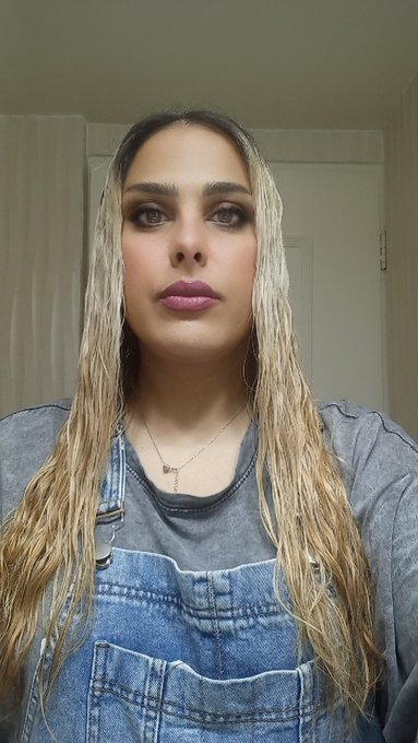 A selfie of a woman with long blonde-streaked hair wearing a denim overall dress and a delicate necklace.