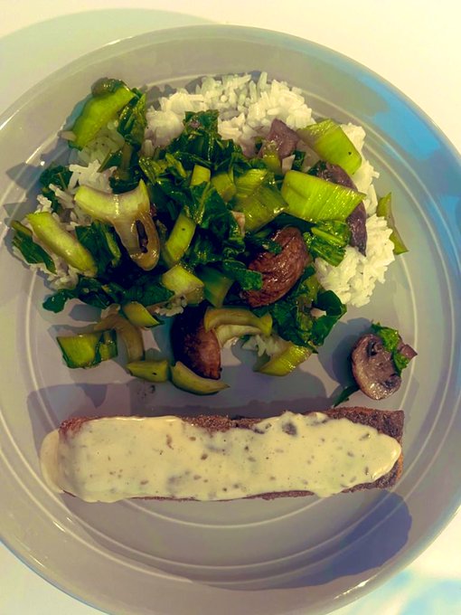 A plate of food featuring a piece of steak topped with creamy white sauce, served alongside white rice mixed with sautéed greens and mushrooms.