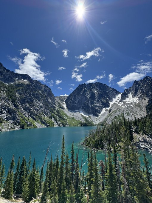 A scenic mountain landscape featuring a turquoise alpine lake surrounded by evergreen trees, with rocky, snow-dusted mountain peaks under a bright blue sky with scattered clouds and a shining sun.