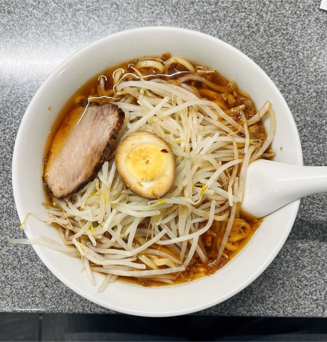 A white bowl filled with ramen sits on a gray surface, containing broth, bean sprouts, chashu pork, and half a boiled egg, accompanied by a white ceramic spoon.