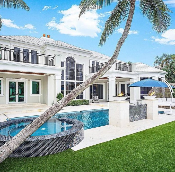 A luxury white two-story Mediterranean-style mansion featuring arched windows, balconies, a swimming pool, an attached spa, an outdoor fireplace, and a leaning palm tree in the foreground on a bright sunny day.