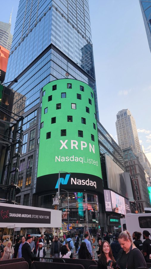 A large digital billboard in Times Square displays "$XRPN" and "Nasdaq Listed" in bright green, alongside the Nasdaq logo, promoting the cryptocurrency's presence on the exchange. The bustling NYC scene below, including the Tamashi Ardows Store, captures the lively atmosphere of Times Square where the XRP branding is prominently featured.